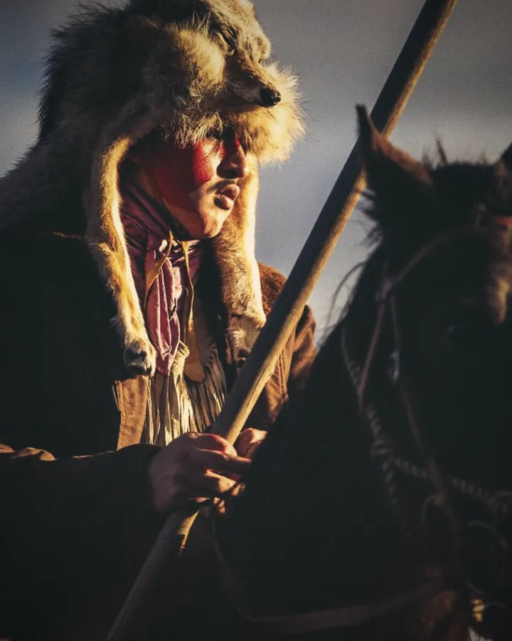A Crow rider on horseback in a wolf-skin headdress, holding a staff