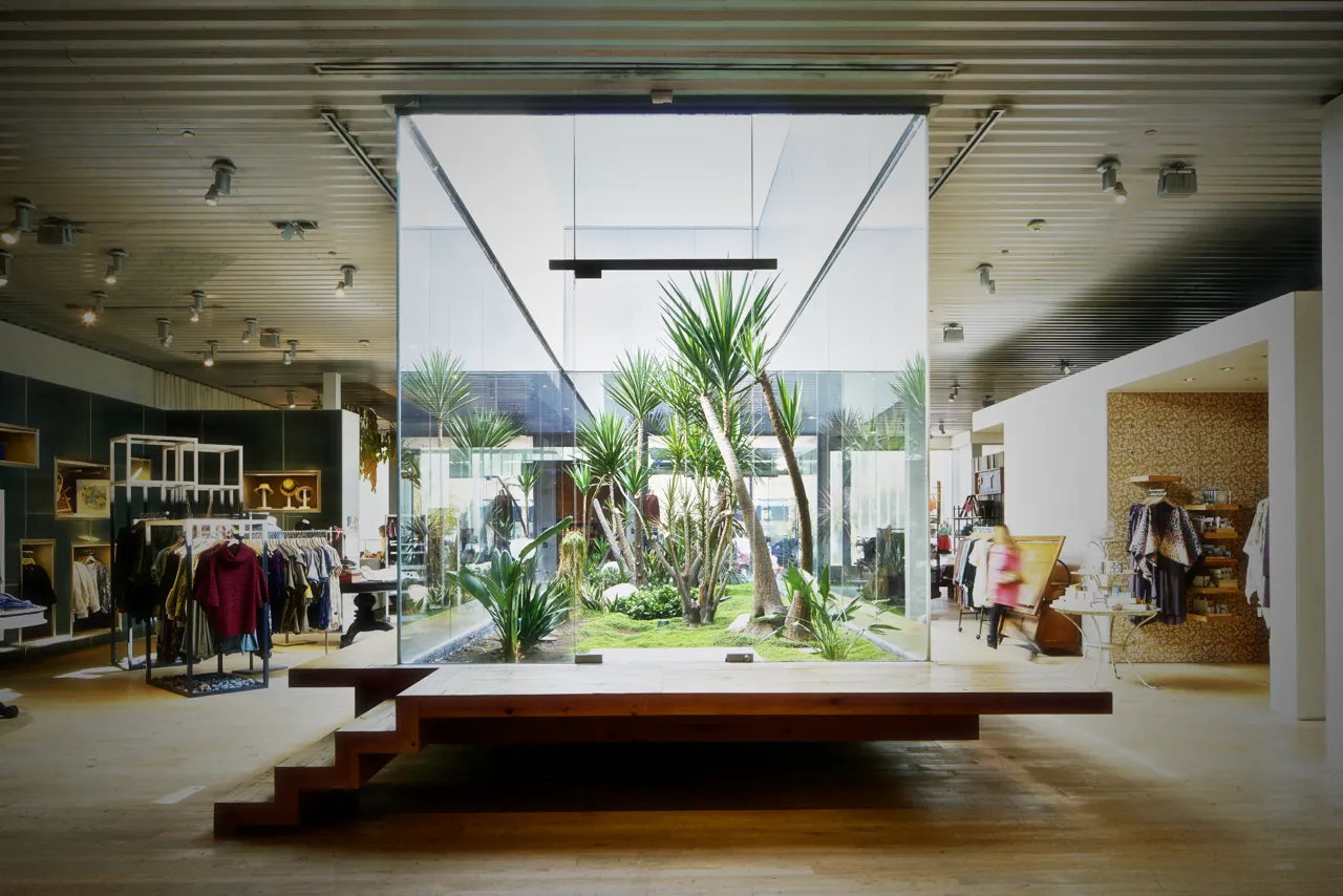 Anthropologie Dos Lagos: a glass room of yucca and grass open to the sky, behind a timber plinth